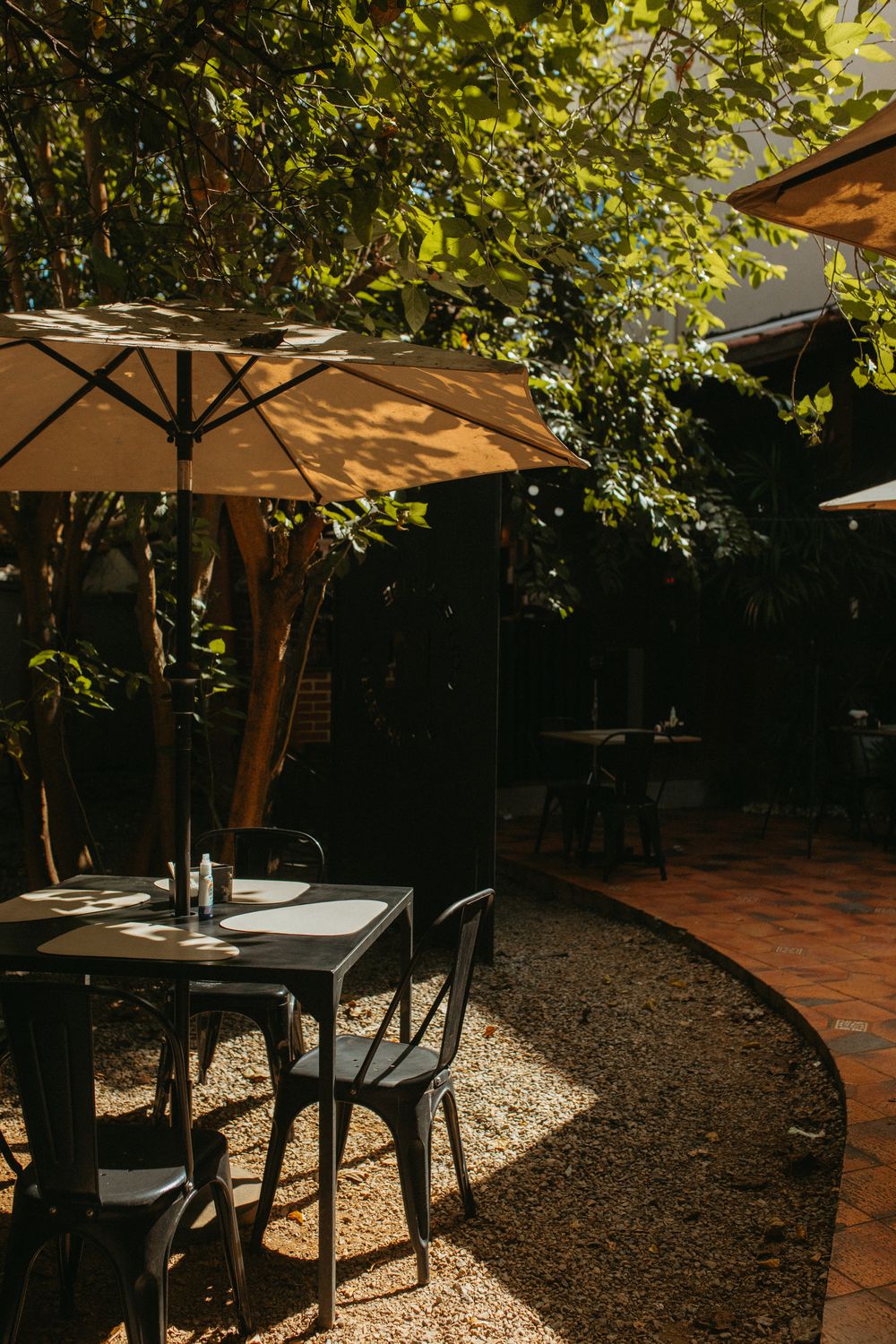 Jardim da Mill com mesa, cadeiras pretas e ombrelone sob as árvores